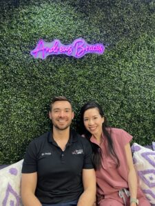 Dr. Philip and Dr. Kat are smiling in front of a grass wall with a neon "Andrews Braces" sign